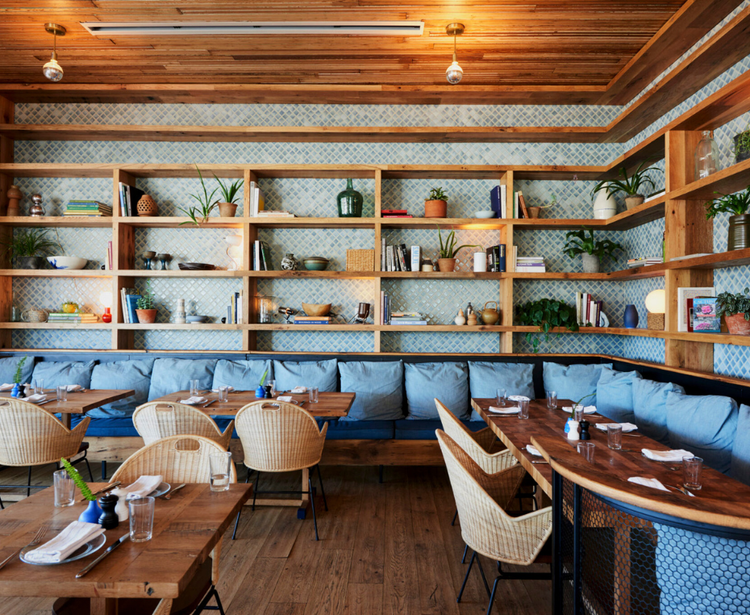 A restaurant interior with wooden tables, blue cushioned seating, and a large shelving unit filled with books, plants, and decorative items.