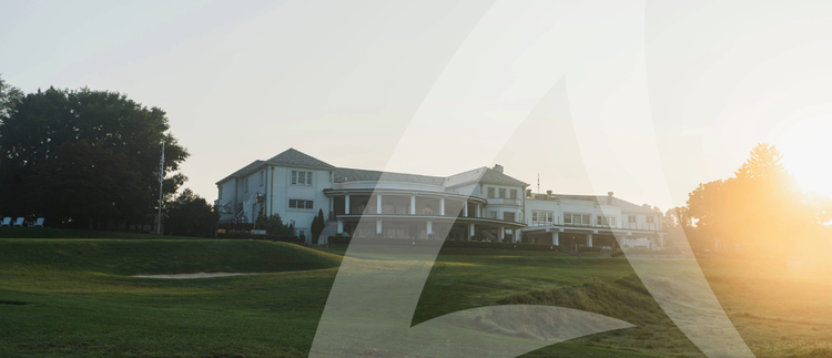 A grand white clubhouse with a porch, columns, and a sloped roof, set on a golf course with a sand bunker, bathed in the warm light of sunrise or sunset.