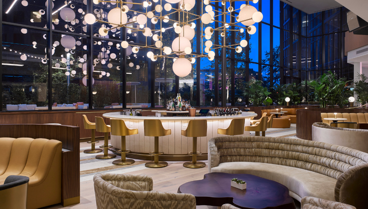 An upscale lounge with a circular bar, gold bar stools, and a modern chandelier, featuring plush seating and floor-to-ceiling windows for an elegant atmosphere.