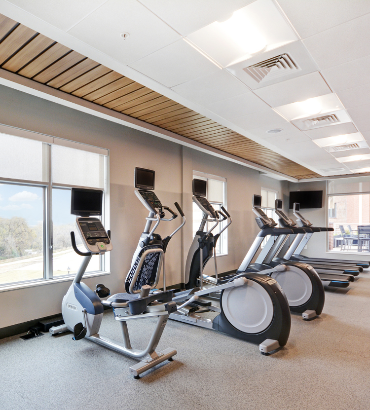 A modern gym with various cardio equipment, including treadmills, stationary bikes, and elliptical machines, with large windows, sleek lighting, and a mounted TV.