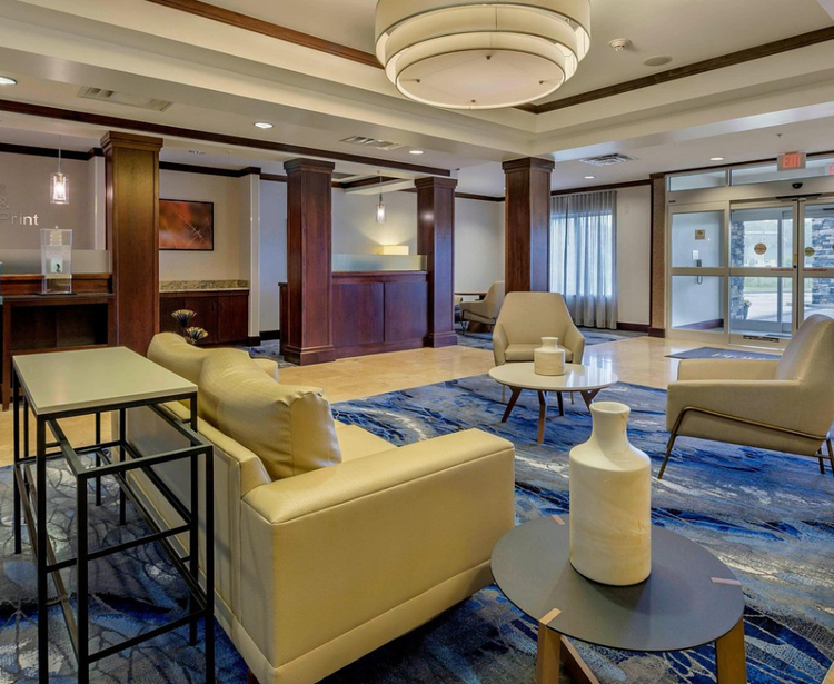 A modern hotel lobby with beige leather seating, round coffee tables, wooden paneling, and blue patterned carpet, creating a warm and welcoming ambiance.
