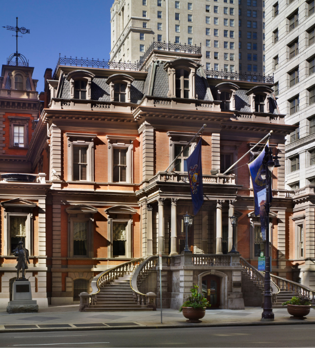 A historic red-brick building with ornate columns, a mansard roof, and arched windows, contrasted by modern skyscrapers in the background.
