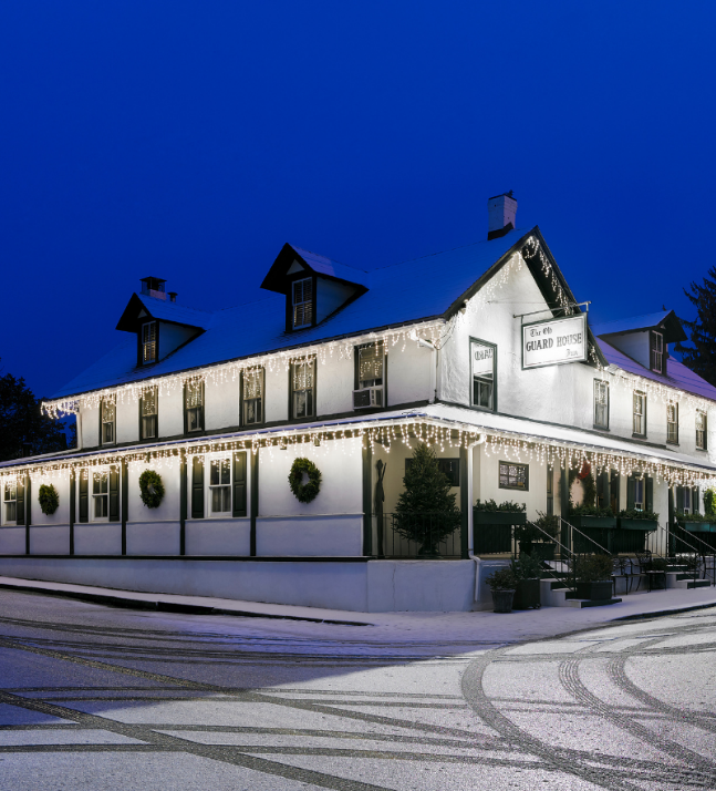 A traditional inn named 'The Old Guard House Inn,' decorated with Christmas lights and wreaths on a snowy evening, creating a festive holiday atmosphere.