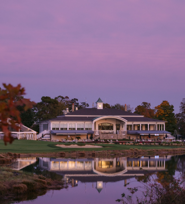 A grand clubhouse with a clock tower, overlooking a reflective body of water, set against a purple-hued sky, with autumn-colored trees in the background.