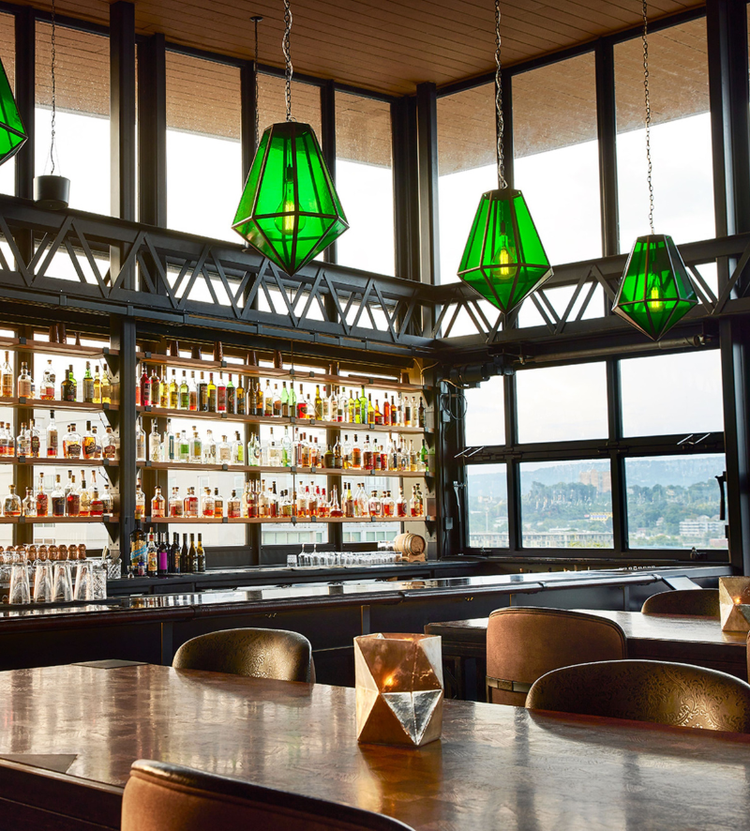 A stylish bar or restaurant interior with large windows, a well-stocked bar, green geometric pendant lights, and tables with decorative candle holders.