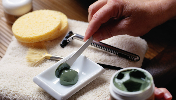 A close-up of a hand holding a spatula with green cream, placing it onto a dish. Surrounding the dish are grooming items like a sponge, brush, and razor on a soft towel.