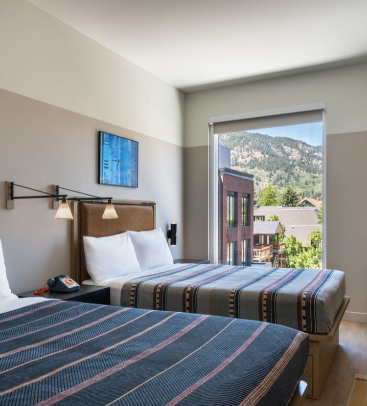 A cozy hotel room with two beds featuring striped blankets, a vintage rotary phone on the nightstand, and a large window showcasing a scenic mountain view.