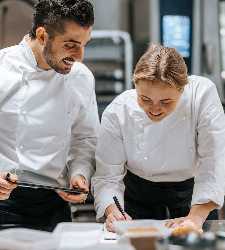 Happy male and female chefs doing bookkeeping in a restaurant kitchen, showcasing Avendra International's culinary and menu support services.
