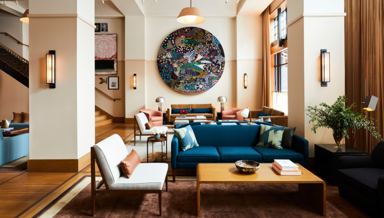 A stylish lounge with a blue sofa, elegant chairs, and a wooden coffee table, featuring a large circular artwork and a staircase leading to another area.