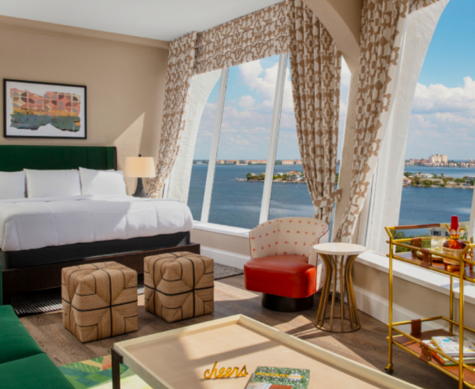 Una elegante habitación de hotel con una vista panorámica desde la ventana, una cama con sábanas blancas y mobiliario moderno, incluidos otomanas y un carrito de bar.