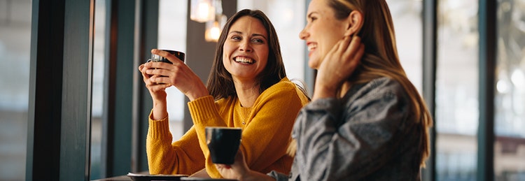 Deux femmes assises à une table, chacune avec une tasse à la main, partageant un moment de complicité et de conversation.