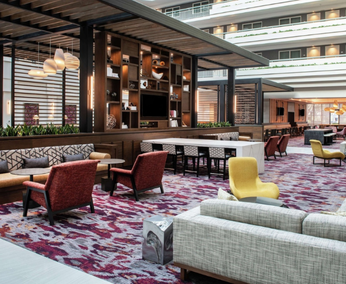 Un hall d'hôtel moderne avec des sièges rouges et jaunes, une moquette à motifs, des étagères en bois et des éléments décoratifs, créant une atmosphère accueillante.