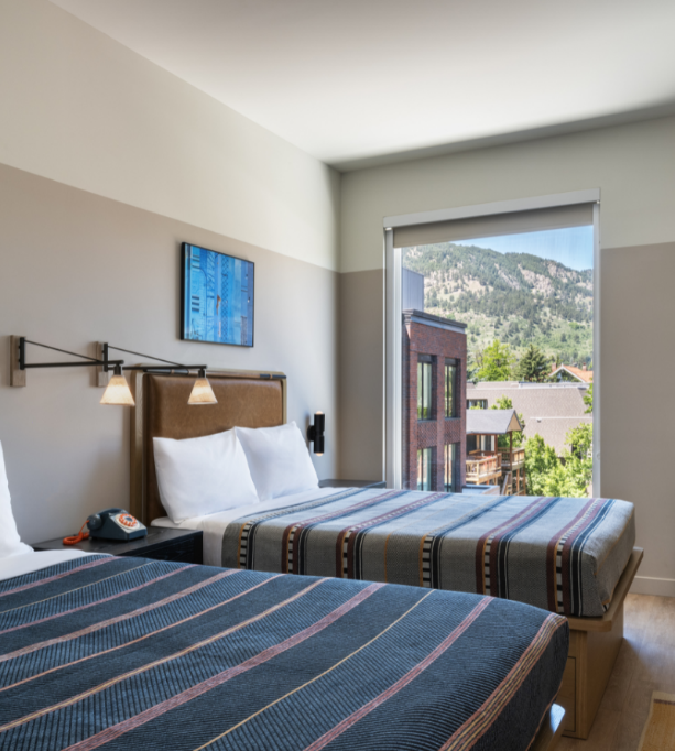 Une chambre d'hôtel confortable avec deux lits aux couvertures rayées, un téléphone à cadran vintage sur la table de nuit et une grande fenêtre offrant une vue pittoresque sur les montagnes.