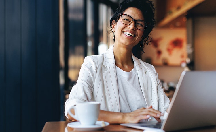 Une personne vêtue d'un blazer blanc et d'une chemise travaille sur un ordinateur portable à la table d'un café, avec une tasse de café à proximité et un arrière-plan chaleureux et confortablement éclairé.