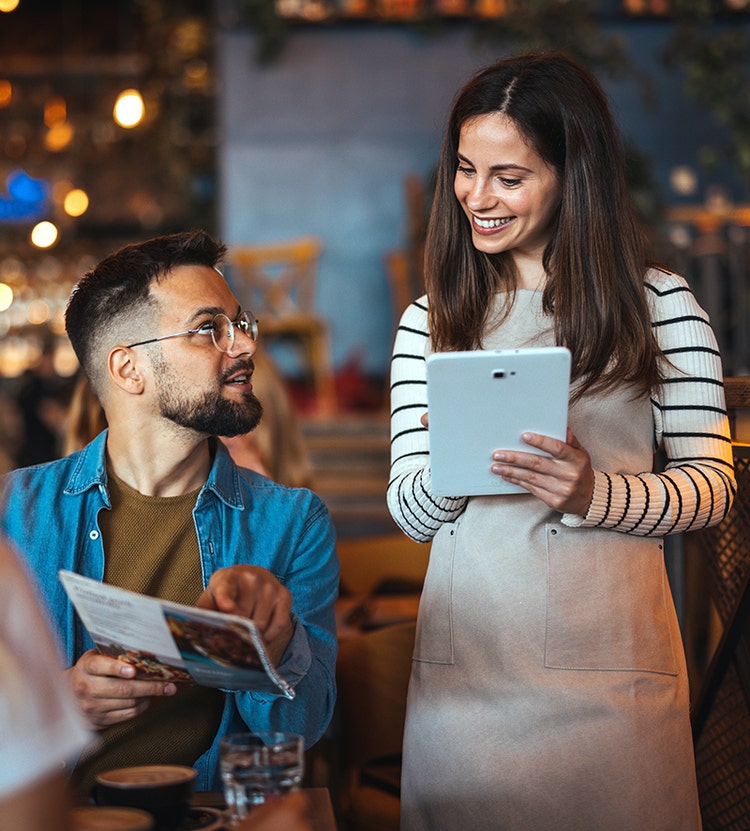 Un client assis à une table dans un restaurant à l'ambiance détendue, tenant un menu et pointant du doigt un plat, tandis qu'un membre du personnel se tient à proximité avec une tablette pour prendre la commande, dans une lumière chaleureuse et un décor animé.