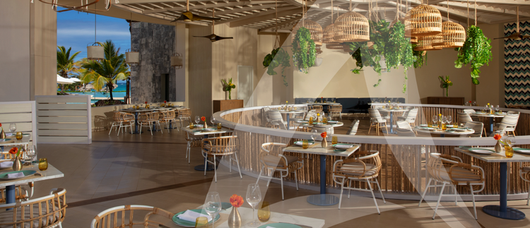 A stylish open-air restaurant with wooden and wicker furniture, warm lighting, and lush greenery, overlooking the ocean for a tropical dining experience.