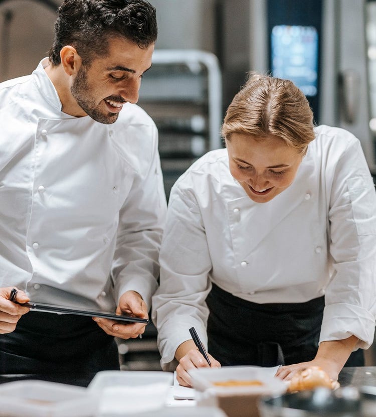 Happy male and female chefs doing bookkeeping in a restaurant kitchen, showcasing Avendra International's culinary and menu support services.