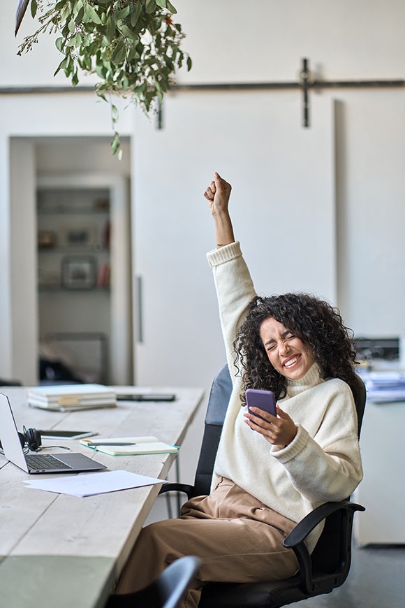 Woman seated at a desk with a phone in one hand and her other hand stretched above her head with an expression of excitement