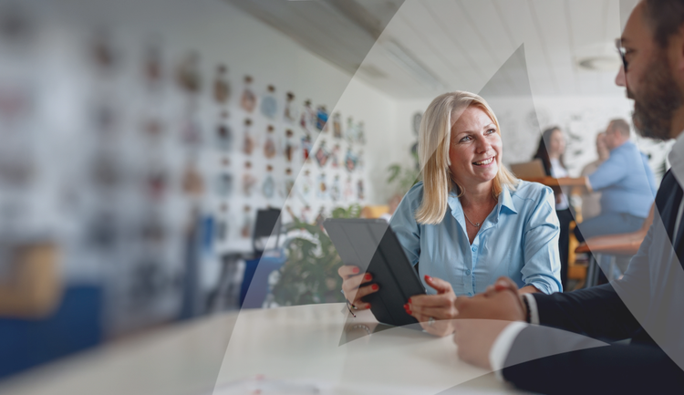Two people seated at a table in a professional setting, engaged in a discussion with a tablet and visible background decor.