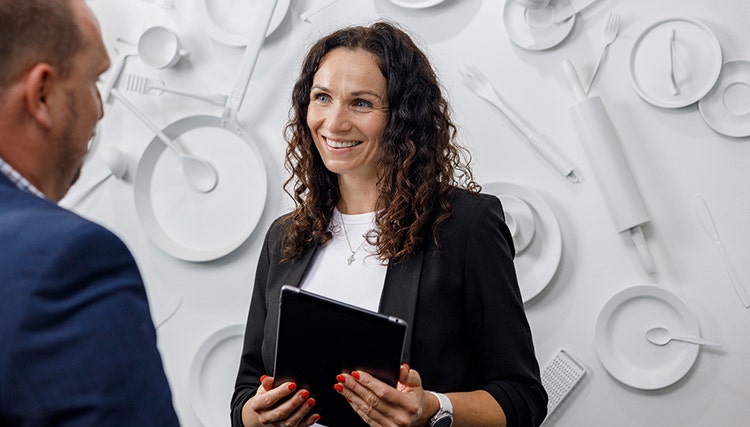 Two people conversing in front of a wall decorated with white kitchen utensils and tableware. One holds a tablet, dressed in a black blazer and white shirt, while the other wears a blue suit jacket.
