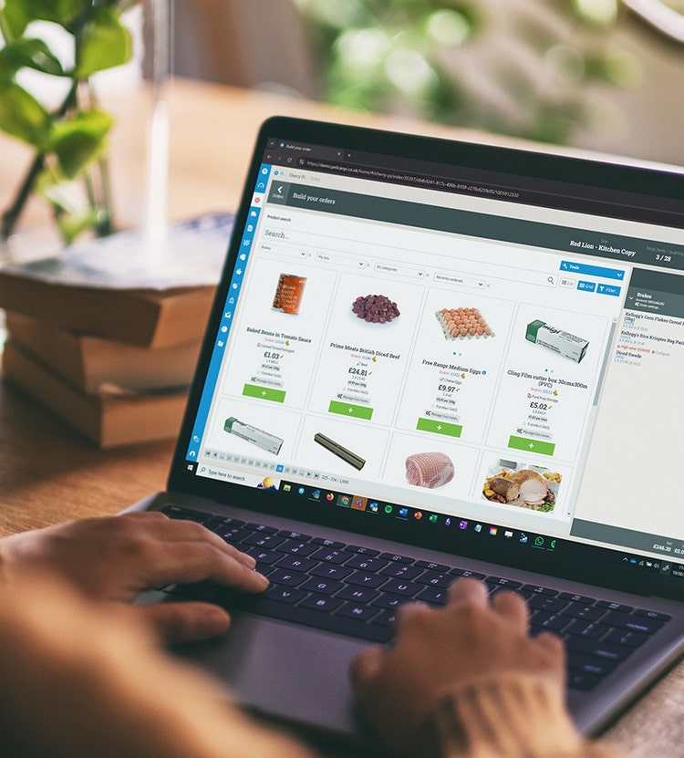 A person shopping for groceries online using a laptop, displaying items like baked beans, beef, eggs, and cling film with prices. The laptop is on a wooden table with books and a plant in the background.