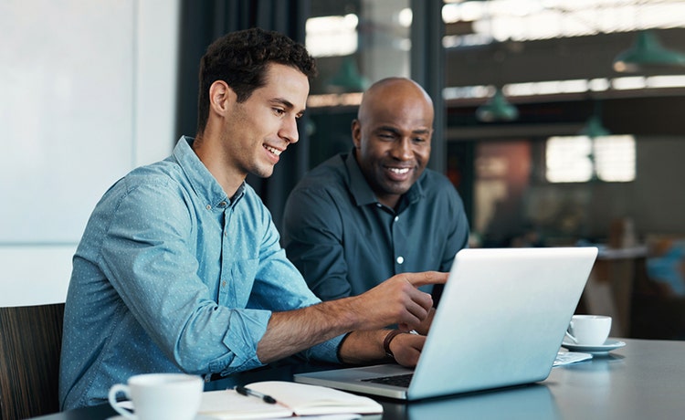 Two people sit at a table reviewing something on a laptop, with coffee cups, a notebook, and a pen on the table, set in a bright and modern workspace.