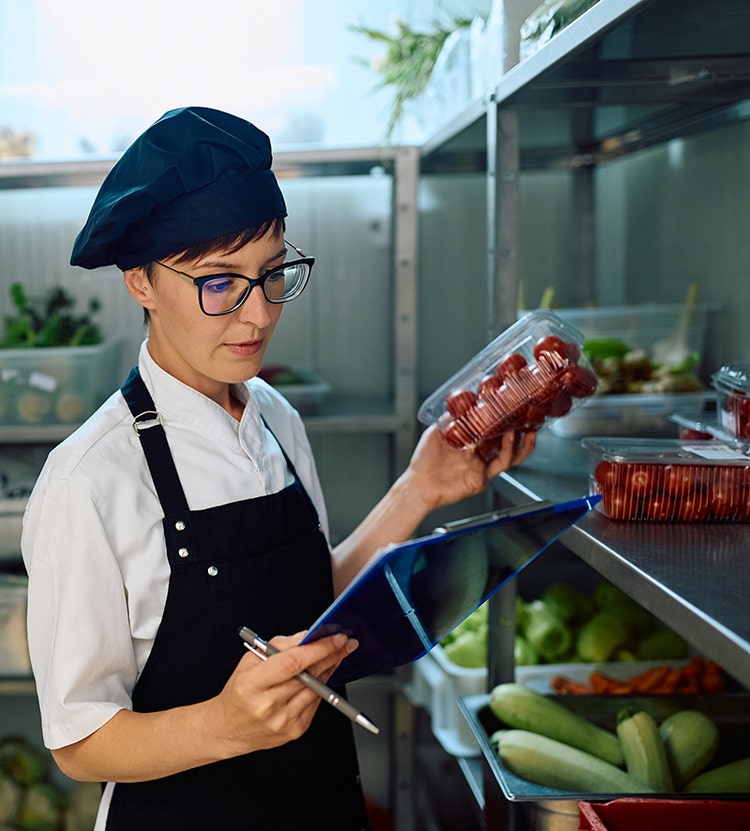 A chef in a white shirt and black apron holding a container of cherry tomatoes and a clipboard with a pen, standing in a pantry with shelves of vegetables like cucumbers and bell peppers.