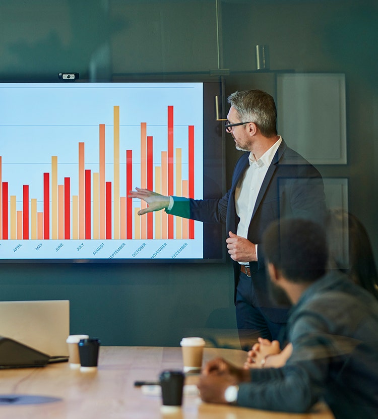 A business meeting in a conference room. A presenter in a suit points at a screen with a bar graph, while three attendees sit at a table with coffee cups and a laptop.