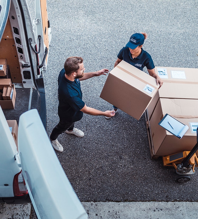 Two people handling cardboard boxes near a delivery van in a warehouse area. One person is in the van, handing a box to the other, who places it on a pallet with a clipboard resting on top.